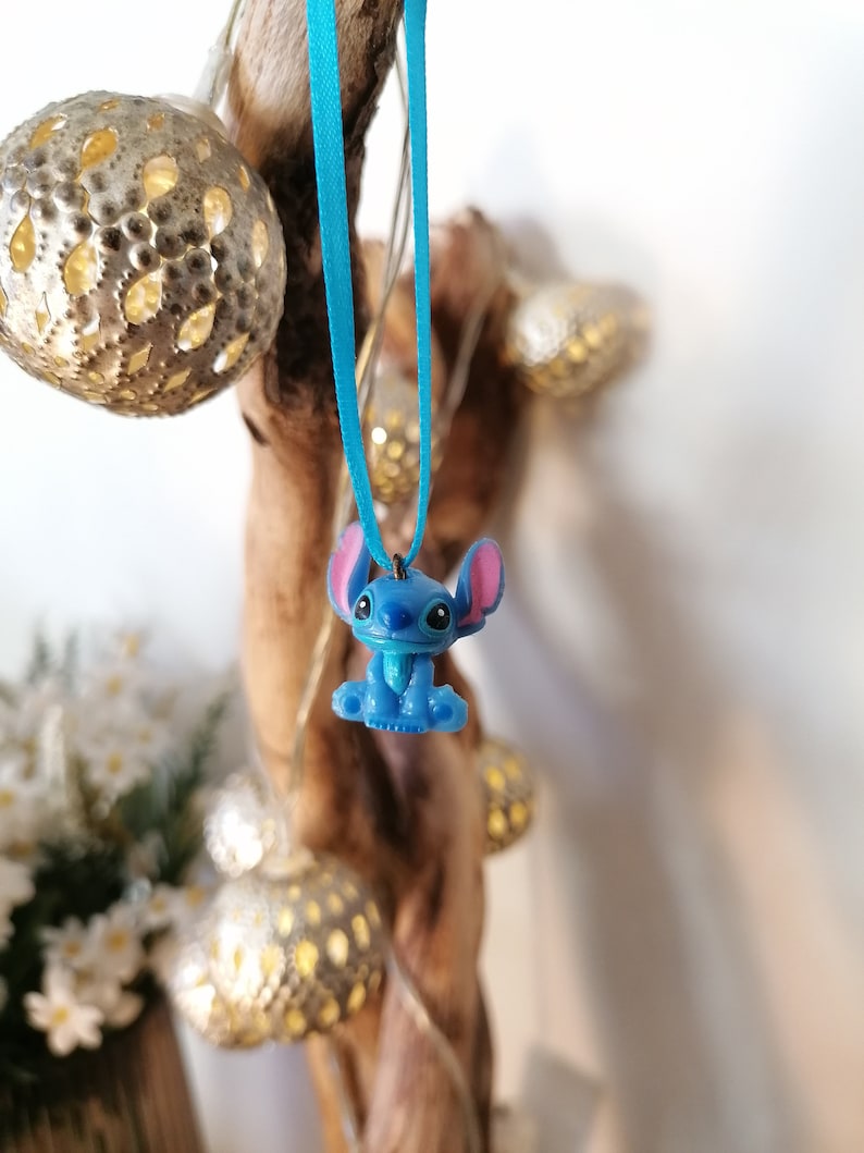 May include: A blue Stitch character charm hanging from a light blue ribbon. The charm is attached to a wooden branch with fairy lights.