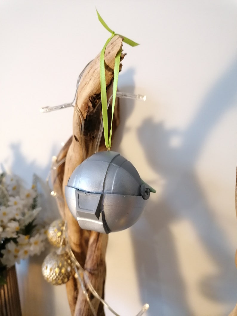 May include: A silver and green Christmas ornament shaped like a spaceship, hanging from a branch with a green ribbon.