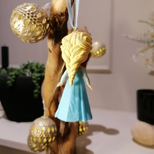 May include: A blue and white ornament of a girl with blonde hair in a braid. The ornament is hanging from a blue ribbon on a wooden branch.