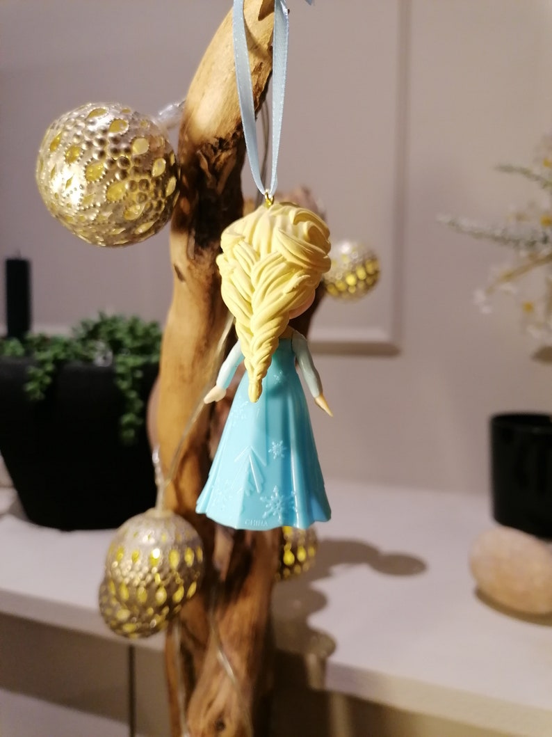 May include: A blue and white ornament of a girl with blonde hair in a braid. The ornament is hanging from a blue ribbon on a wooden branch.