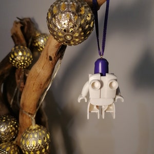 May include: White Lego minifigure ornament with a purple head and a white body. The ornament is hanging from a purple ribbon on a branch with gold and white decorative balls.