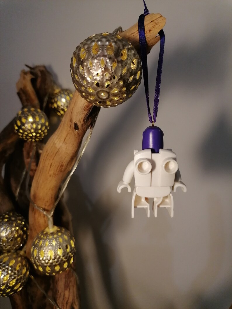 May include: White Lego minifigure ornament with a purple head and a white body. The ornament is hanging from a purple ribbon on a branch with gold and white decorative balls.