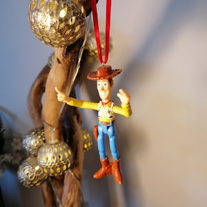 May include: A red string hangs from a wooden branch with a plastic toy figurine of Woody from Toy Story. Woody is wearing his signature cowboy hat, vest, and jeans. He is holding his right arm up in the air, as if he is waving.