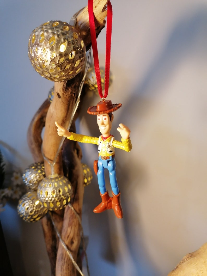 May include: A red string hangs from a wooden branch with a plastic toy figurine of Woody from Toy Story. Woody is wearing his signature cowboy hat, vest, and jeans. He is holding his right arm up in the air, as if he is waving.