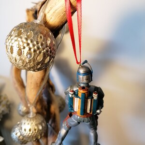 May include: A blue and gray Star Wars Boba Fett Christmas ornament with a red ribbon hanging from a wooden branch.