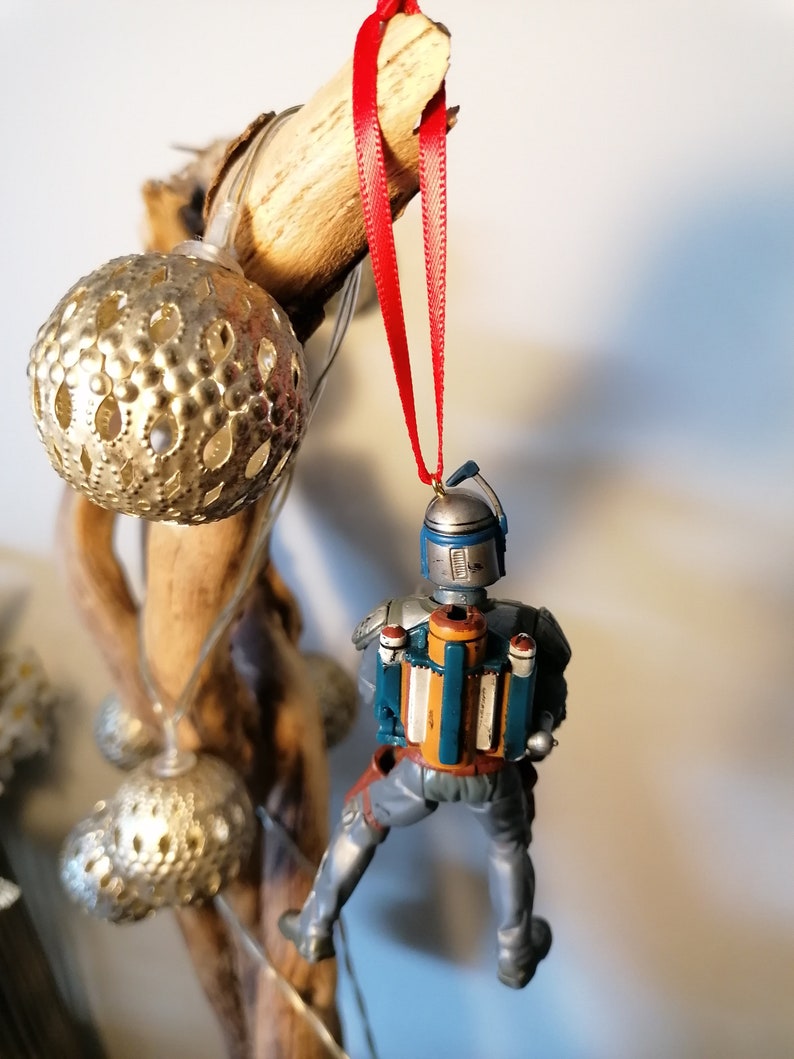 May include: A blue and gray Star Wars Boba Fett Christmas ornament with a red ribbon hanging from a wooden branch.