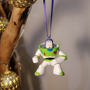 May include: A green and white plastic figurine of Buzz Lightyear from the Toy Story movie franchise. The figurine is hanging from a purple ribbon and has a white background.