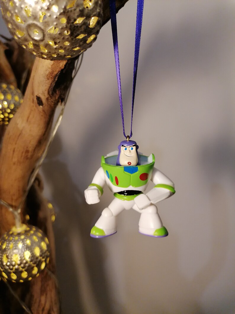 May include: A green and white plastic figurine of Buzz Lightyear from the Toy Story movie franchise. The figurine is hanging from a purple ribbon and has a white background.