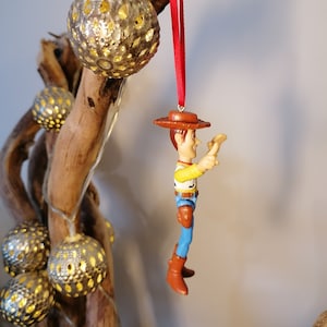 May include: A red string ornament with a plastic figurine of Woody from Toy Story. Woody is wearing his signature cowboy hat, vest, and jeans. He is holding his arm out, as if he is reaching for something.