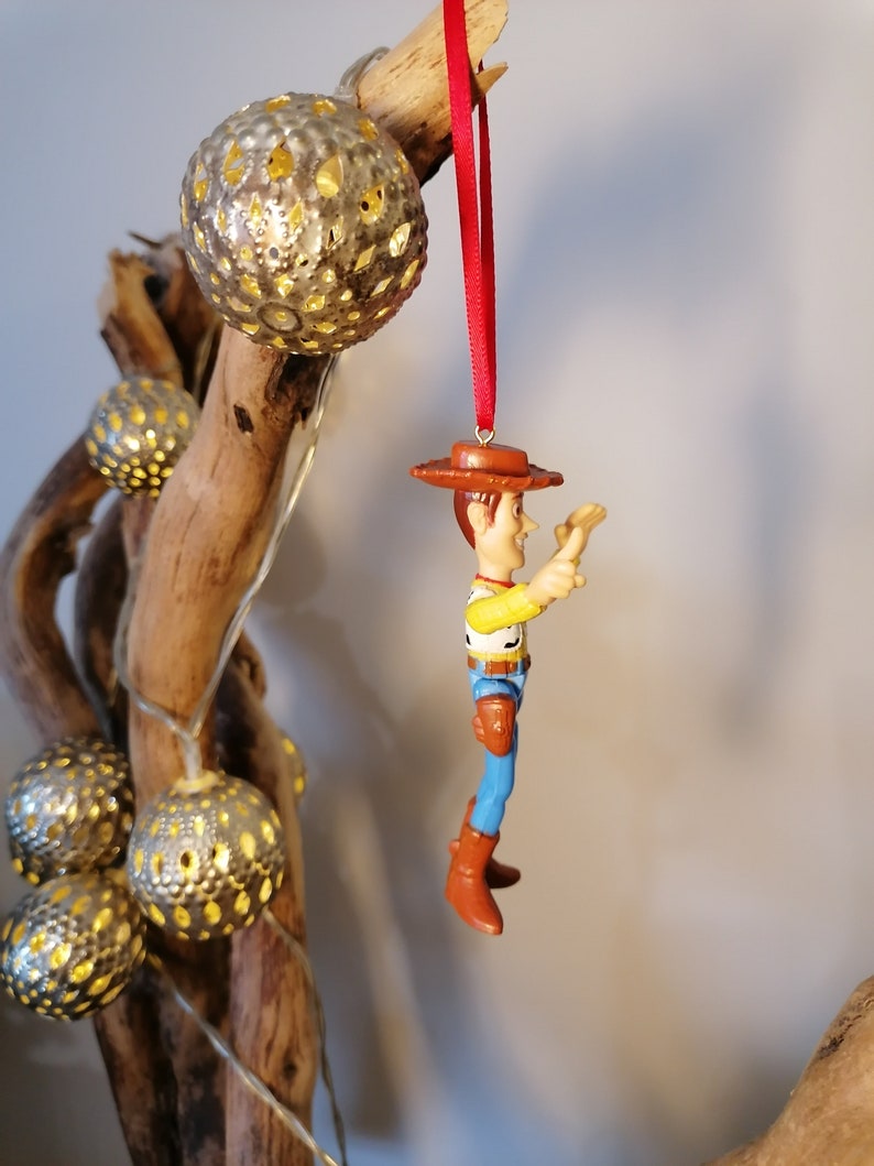 May include: A red string ornament with a plastic figurine of Woody from Toy Story. Woody is wearing his signature cowboy hat, vest, and jeans. He is holding his arm out, as if he is reaching for something.