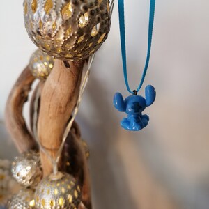May include: A blue Stitch character charm hangs from a turquoise blue ribbon necklace. The necklace is hanging from a branch with decorative lights.