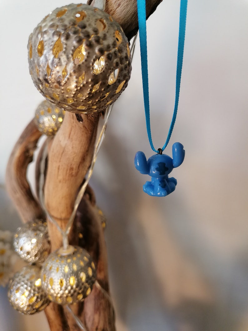 May include: A blue Stitch character charm hangs from a turquoise blue ribbon necklace. The necklace is hanging from a branch with decorative lights.