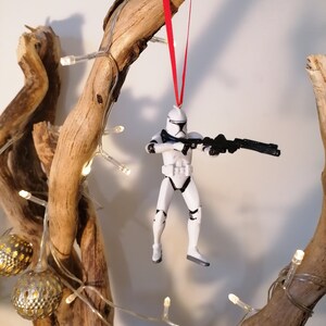 Puede incluir: Un adorno de figura de acci&oacute;n de Stormtrooper blanco colgado de una rama con una cinta roja. La figura sostiene un rifle bl&aacute;ster y lleva un casco y una armadura blancos.