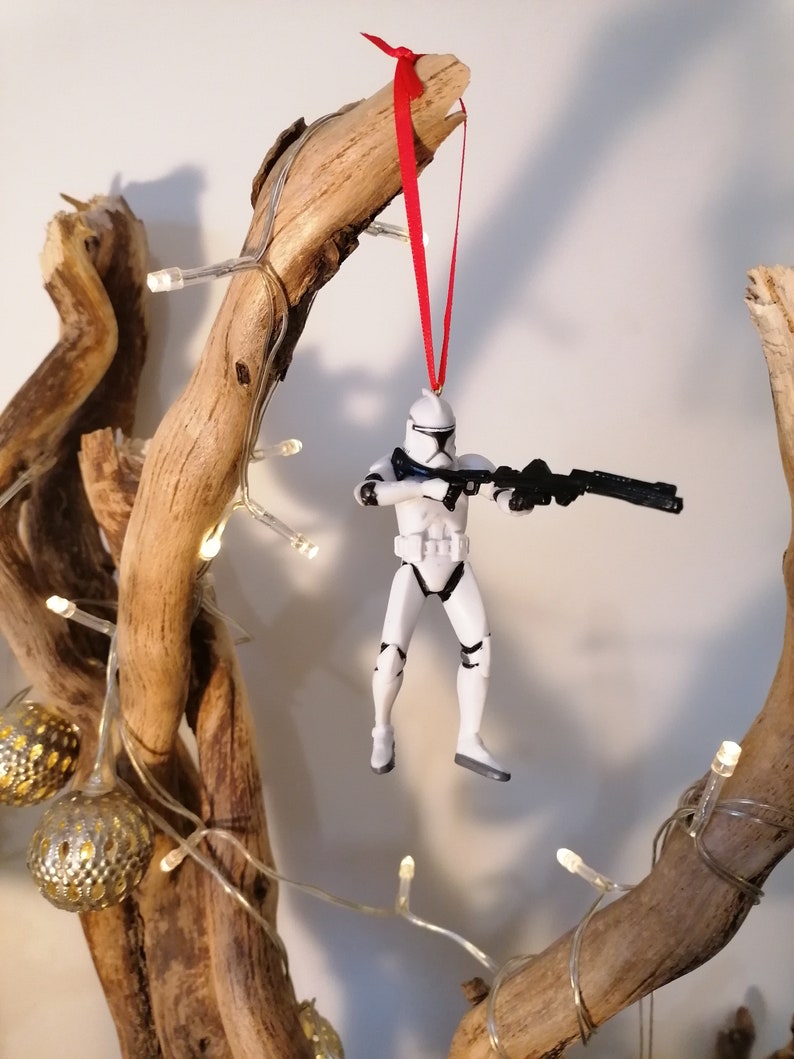Puede incluir: Un adorno de figura de acci&oacute;n de Stormtrooper blanco colgado de una rama con una cinta roja. La figura sostiene un rifle bl&aacute;ster y lleva un casco y una armadura blancos.