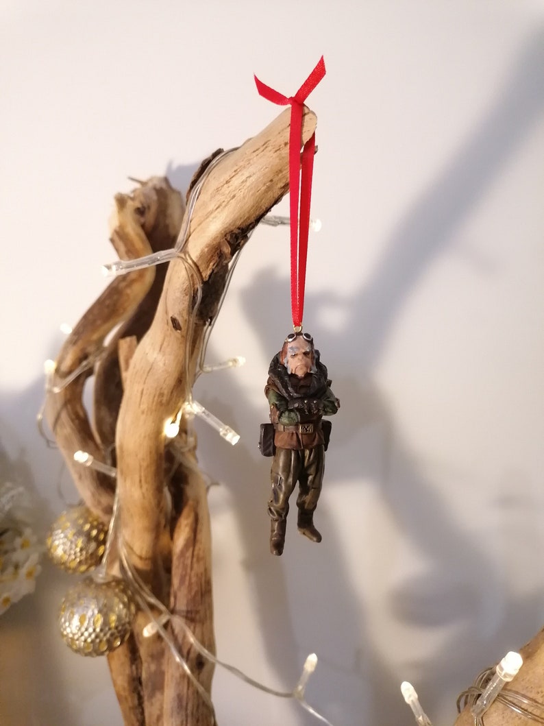 K&ouml;nnte beinhalten: Detaillierter Weihnachtsschmuck, der eine Milit&auml;rfigur in Tarnkleidung darstellt, die an einem roten Band aufgeh&auml;ngt ist. Der Schmuck hat ein braunes und gr&uuml;nes Farbschema mit aufwendigen Details an Uniform und Zubeh&ouml;r. Der Schmuck h&auml;ngt an einem St&uuml;ck Treibholz.