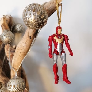 Pode incluir: Um ornamento de figura de a&ccedil;&atilde;o do Homem de Ferro vermelho e prateado pendurado em um galho de madeira &agrave; deriva decorado com bolas douradas brilhantes.