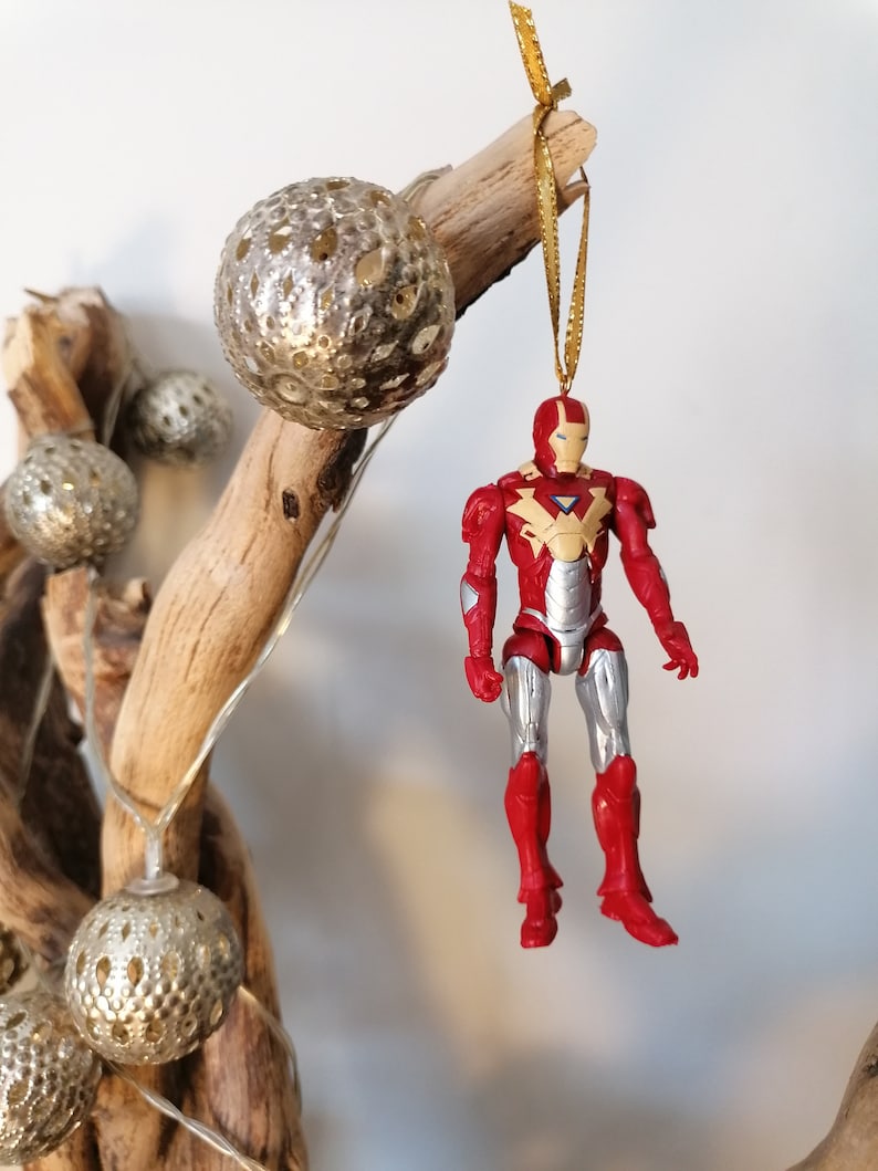 Pode incluir: Um ornamento de figura de a&ccedil;&atilde;o do Homem de Ferro vermelho e prateado pendurado em um galho de madeira &agrave; deriva decorado com bolas douradas brilhantes.
