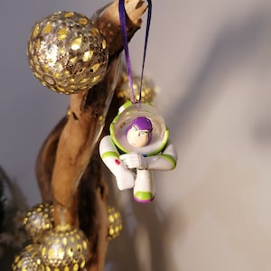 Puede incluir: Un adorno de pl&aacute;stico blanco y verde de Buzz Lightyear de la franquicia Toy Story. El adorno est&aacute; colgado de una cinta morada en una rama con una cadena de luces.