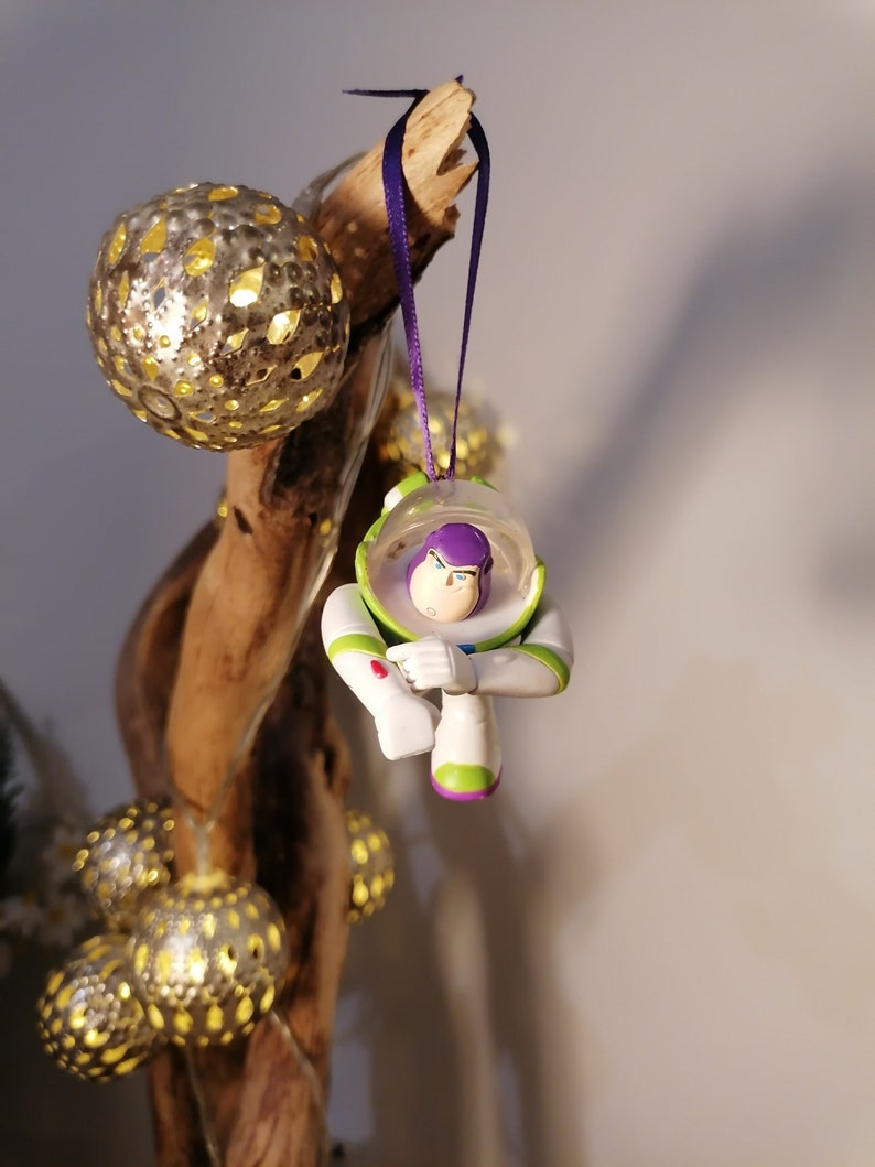 Puede incluir: Un adorno de pl&aacute;stico blanco y verde de Buzz Lightyear de la franquicia Toy Story. El adorno est&aacute; colgado de una cinta morada en una rama con una cadena de luces.