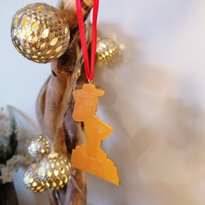 May include: A gold-colored Christmas ornament shaped like a person standing on a platform. The ornament is hanging from a red ribbon on a branch with gold-colored lights.