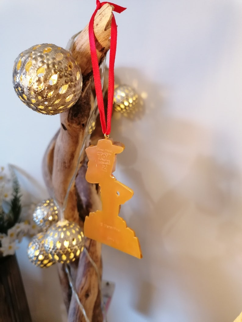 May include: A gold-colored Christmas ornament shaped like a person standing on a platform. The ornament is hanging from a red ribbon on a branch with gold-colored lights.