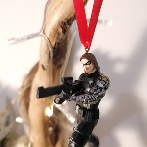 May include: A black and gray action figure of a superhero holding a gun. The figure is wearing a black and gray costume with a white star on the shoulder. The figure is hanging from a red ribbon.