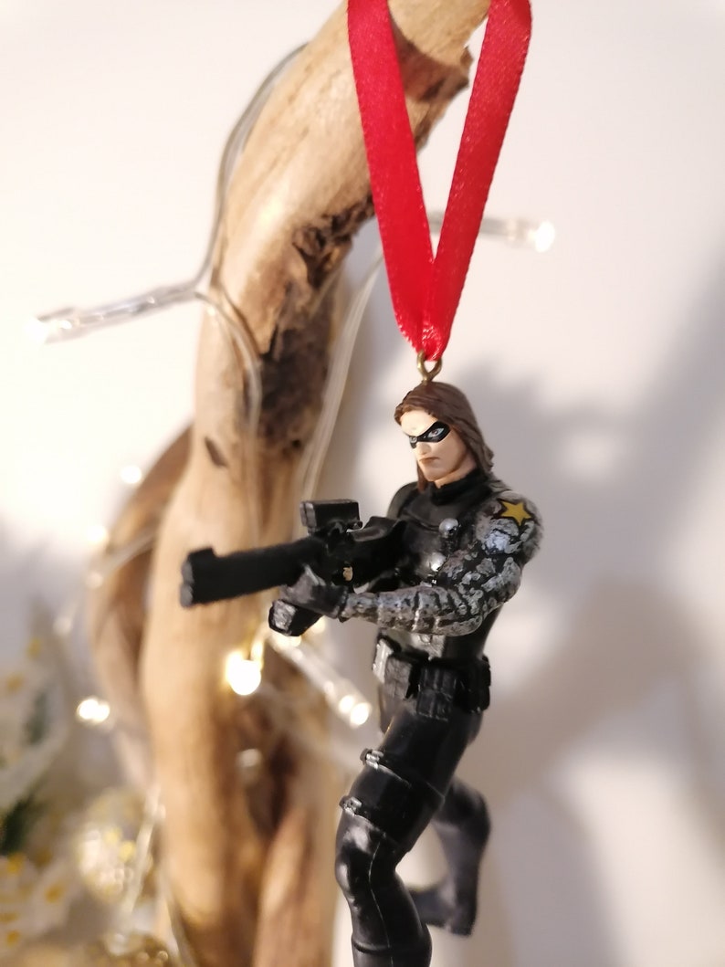 May include: A black and gray action figure of a superhero holding a gun. The figure is wearing a black and gray costume with a white star on the shoulder. The figure is hanging from a red ribbon.