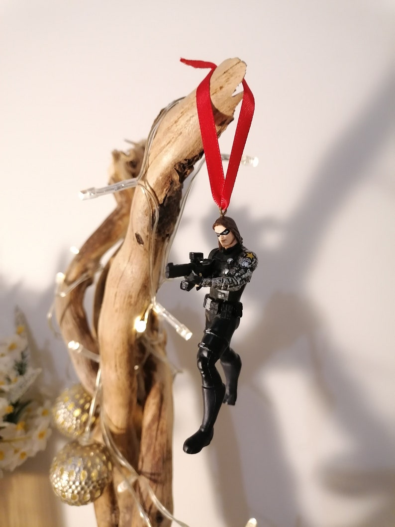 May include: A black and gray action figure of a superhero holding a gun, hanging from a red ribbon on a piece of driftwood.