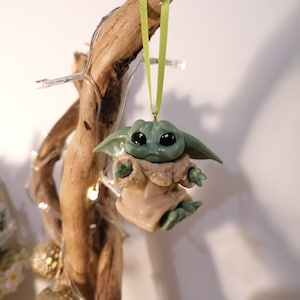 May include: A green and brown Christmas ornament shaped like a character from the Star Wars franchise. The ornament is hanging from a green ribbon on a branch.
