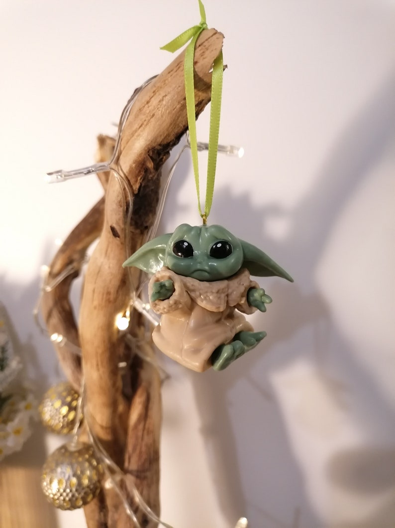 May include: A green and brown Christmas ornament shaped like a character from the Star Wars franchise. The ornament is hanging from a green ribbon on a branch.