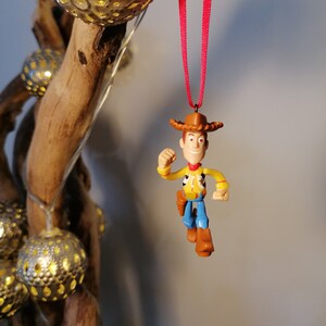 May include: A red string hangs from a wooden branch with a plastic toy figurine of Woody from Toy Story. Woody is wearing his signature cowboy hat, vest, and jeans. He is holding his arm out in a gesture of running.