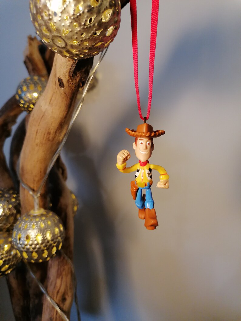 May include: A red string hangs from a wooden branch with a plastic toy figurine of Woody from Toy Story. Woody is wearing his signature cowboy hat, vest, and jeans. He is holding his arm out in a gesture of running.