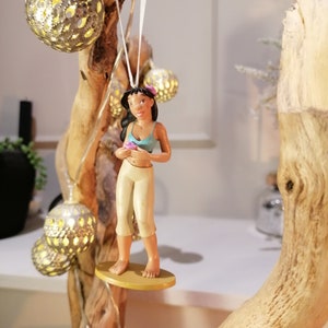 May include: A brown-skinned cartoon character with black hair and a blue top is holding a pink flower. The character is standing on a surfboard with a gold base. The character is hanging from a white string.
