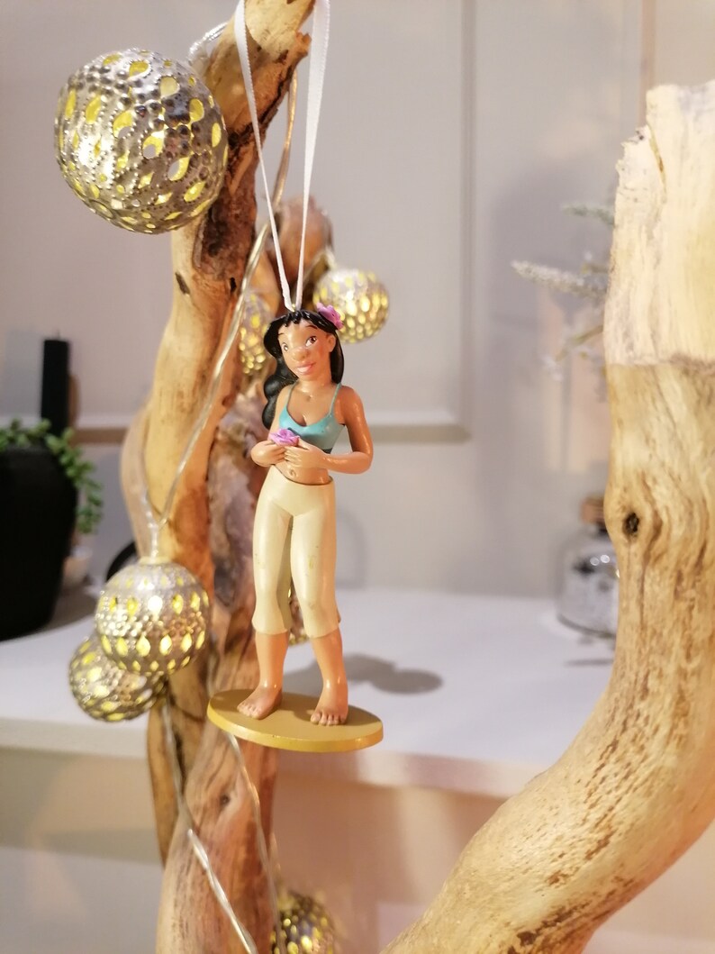 May include: A brown-skinned cartoon character with black hair and a blue top is holding a pink flower. The character is standing on a surfboard with a gold base. The character is hanging from a white string.