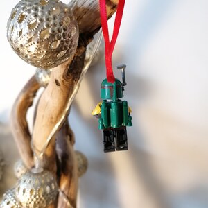 May include: A green and yellow Lego Star Wars Boba Fett minifigure ornament hanging from a red ribbon on a piece of driftwood.
