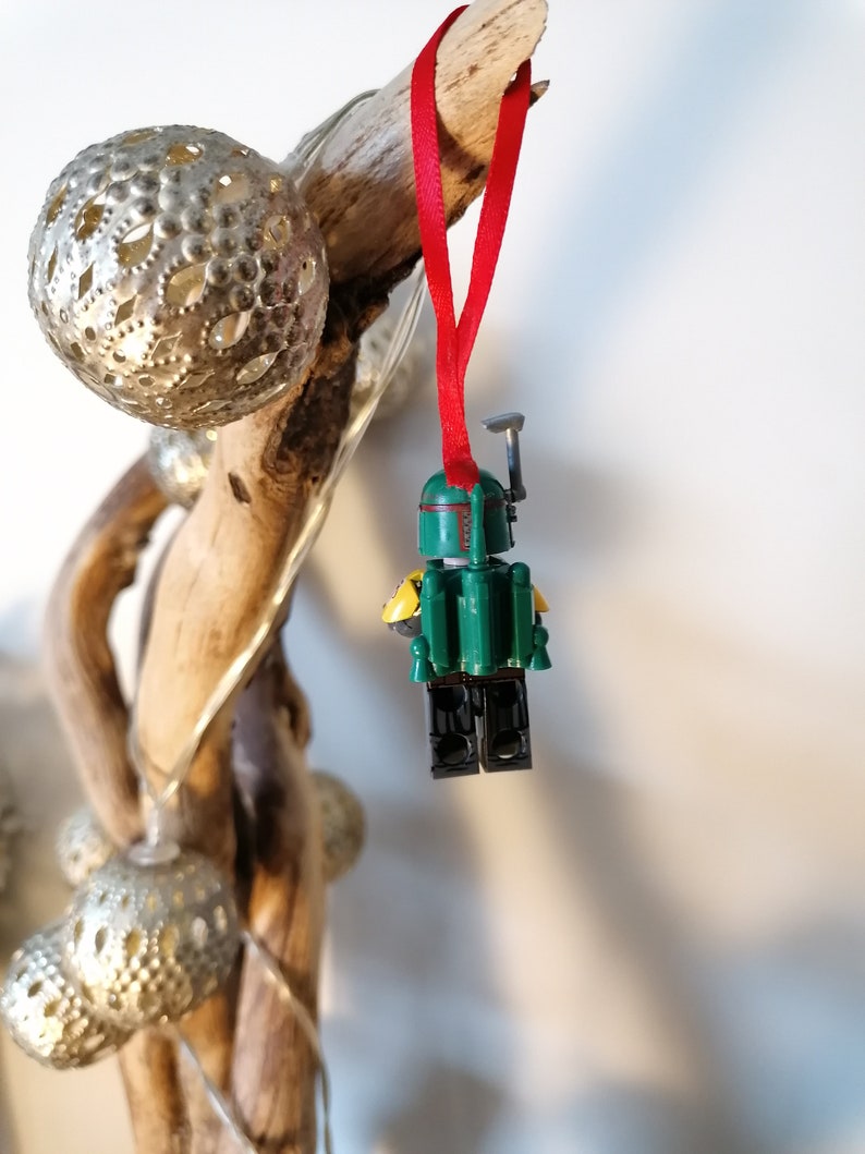 May include: A green and yellow Lego Star Wars Boba Fett minifigure ornament hanging from a red ribbon on a piece of driftwood.