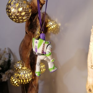 May include: A white and purple plastic ornament of Buzz Lightyear from the Toy Story movie franchise. The ornament is hanging from a purple ribbon on a wooden branch.