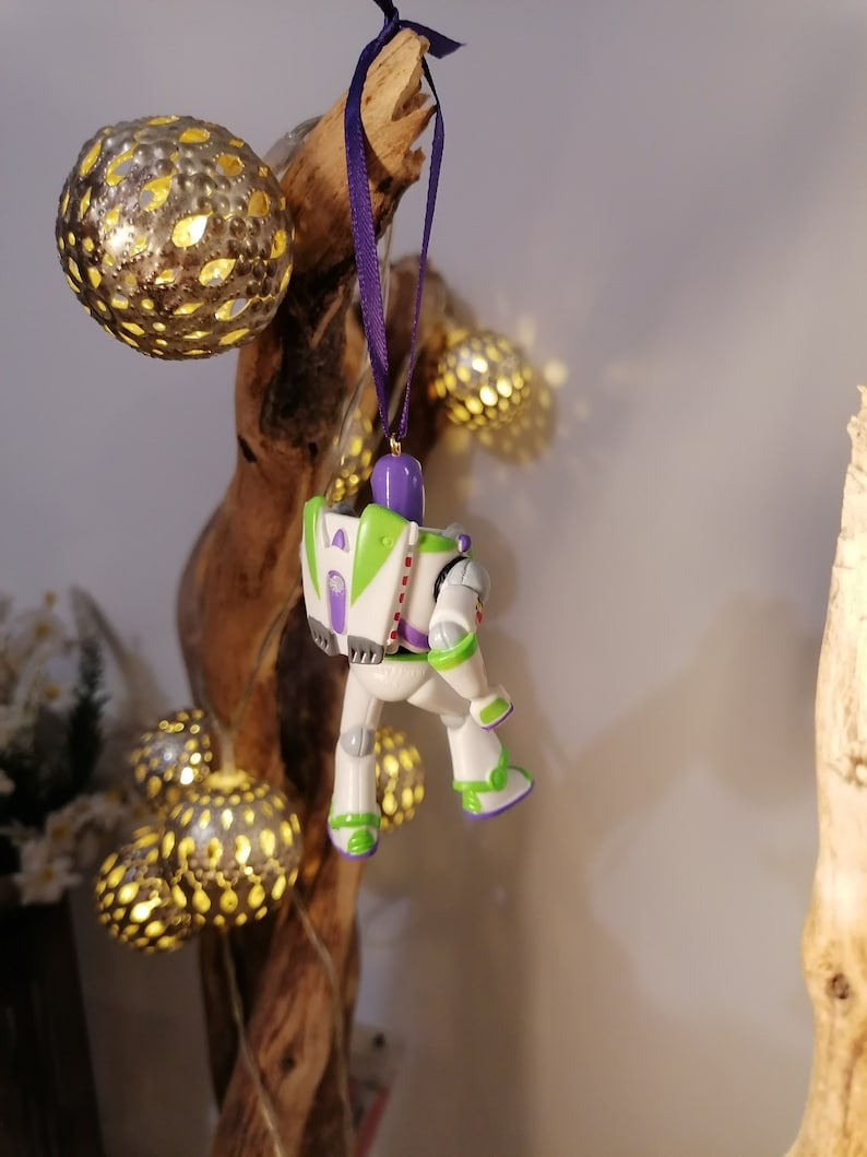 May include: A white and purple plastic ornament of Buzz Lightyear from the Toy Story movie franchise. The ornament is hanging from a purple ribbon on a wooden branch.
