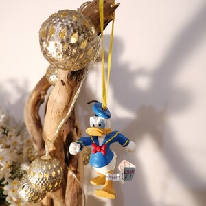 以下が含まれることがあります： 黄色のリボンに青いプラスチック製のドナルドダックのクリスマスオーナメントがぶら下がっています。オーナメントは白い蝶ネクタイをしています。オーナメントは、電飾がついた枝にぶら下がっています。