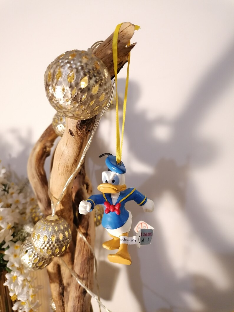 以下が含まれることがあります： 黄色のリボンに青いプラスチック製のドナルドダックのクリスマスオーナメントがぶら下がっています。オーナメントは白い蝶ネクタイをしています。オーナメントは、電飾がついた枝にぶら下がっています。