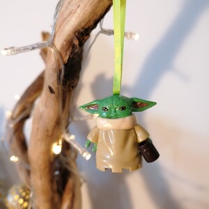 May include: A green and brown Yoda ornament with a green face and large ears. The ornament is holding a brown cup in its right hand. The ornament is hanging from a green ribbon on a branch.