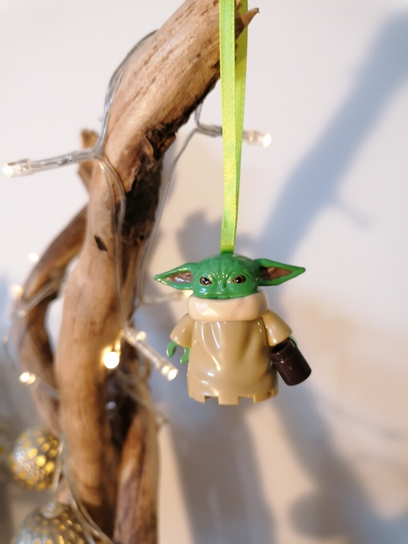 May include: A green and brown Yoda ornament with a green face and large ears. The ornament is holding a brown cup in its right hand. The ornament is hanging from a green ribbon on a branch.