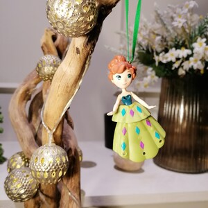 May include: A green and purple dress ornament with a light brown haired doll wearing a blue top. The ornament is hanging from a green string on a wooden branch with gold decorative balls.