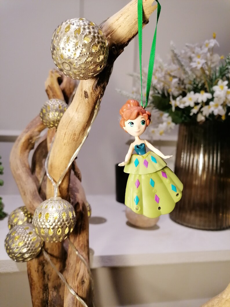 May include: A green and purple dress ornament with a light brown haired doll wearing a blue top. The ornament is hanging from a green string on a wooden branch with gold decorative balls.