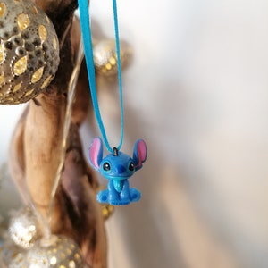 May include: Blue Stitch character charm hanging from a turquoise ribbon. The charm is a popular Disney character with large ears and a friendly expression.