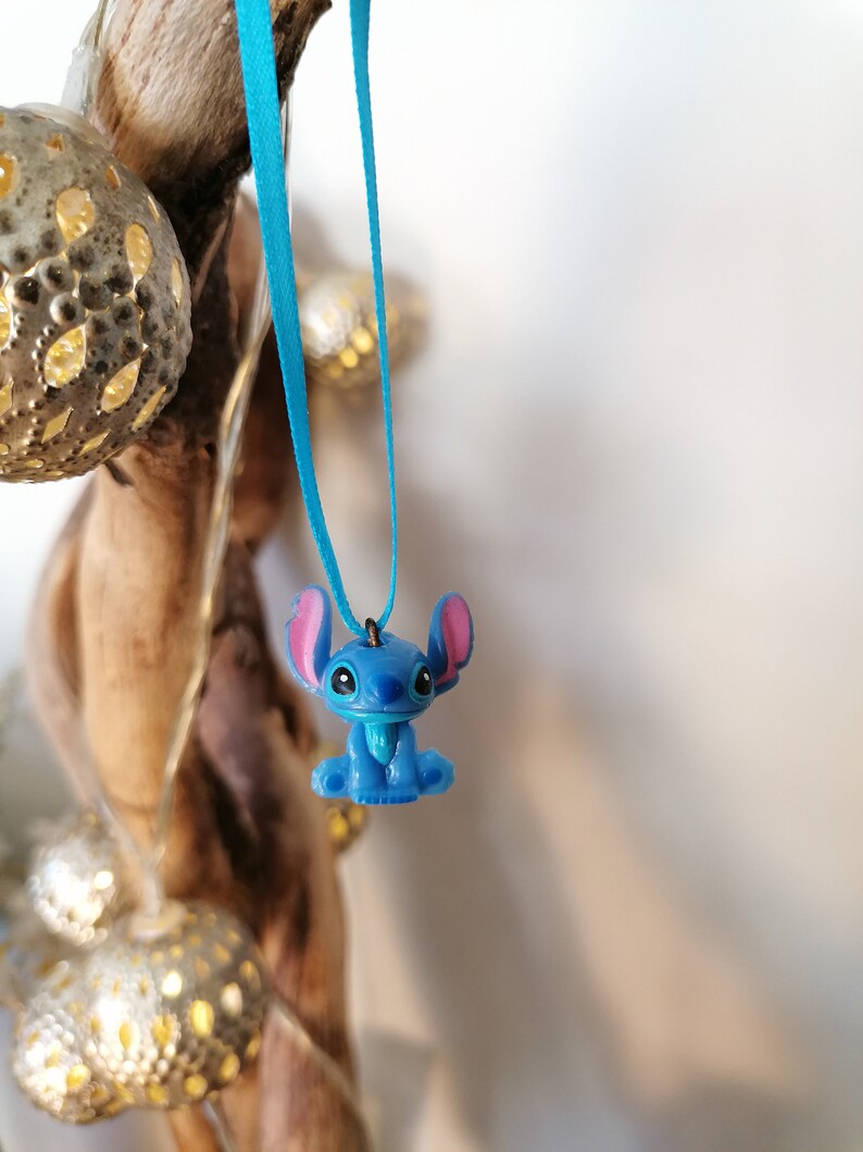 May include: Blue Stitch character charm hanging from a turquoise ribbon. The charm is a popular Disney character with large ears and a friendly expression.