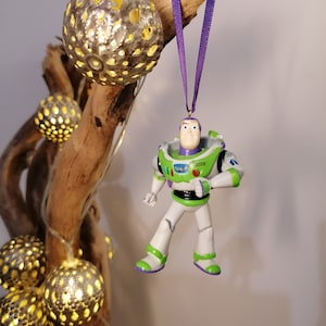 Puede incluir: Una figura de juguete de plástico verde y blanco de Buzz Lightyear de la franquicia de películas Toy Story colgando de una cinta morada. La figura lleva un uniforme de ranger espacial con un cinturón negro y un botón rojo en el pecho. La figura está colgando de una rama con luces de cadena.