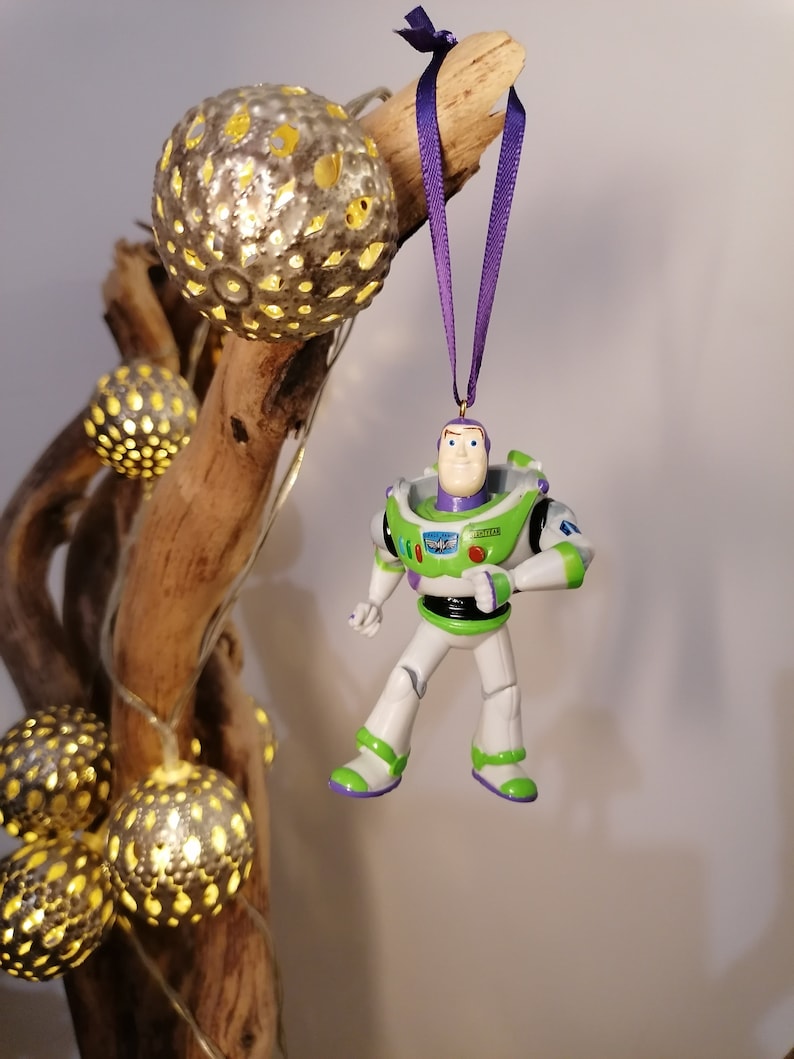 May include: A green and white plastic toy figurine of Buzz Lightyear from the Toy Story movie franchise hanging from a purple ribbon. The figurine is wearing a space ranger uniform with a black belt and a red button on his chest. The figurine is hanging from a branch with string lights.