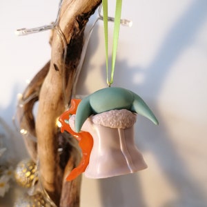 May include: A green and purple Christmas bauble shaped like a character from the Star Wars franchise. The bauble is wearing a white fur coat and has a green head with large ears. The bauble is hanging from a green ribbon.