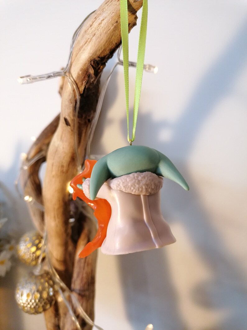 May include: A green and purple Christmas bauble shaped like a character from the Star Wars franchise. The bauble is wearing a white fur coat and has a green head with large ears. The bauble is hanging from a green ribbon.